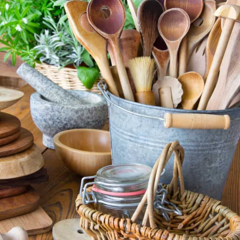 Bucket of wooden utensils on a table with other bowls, baskets, and cooking supplies.