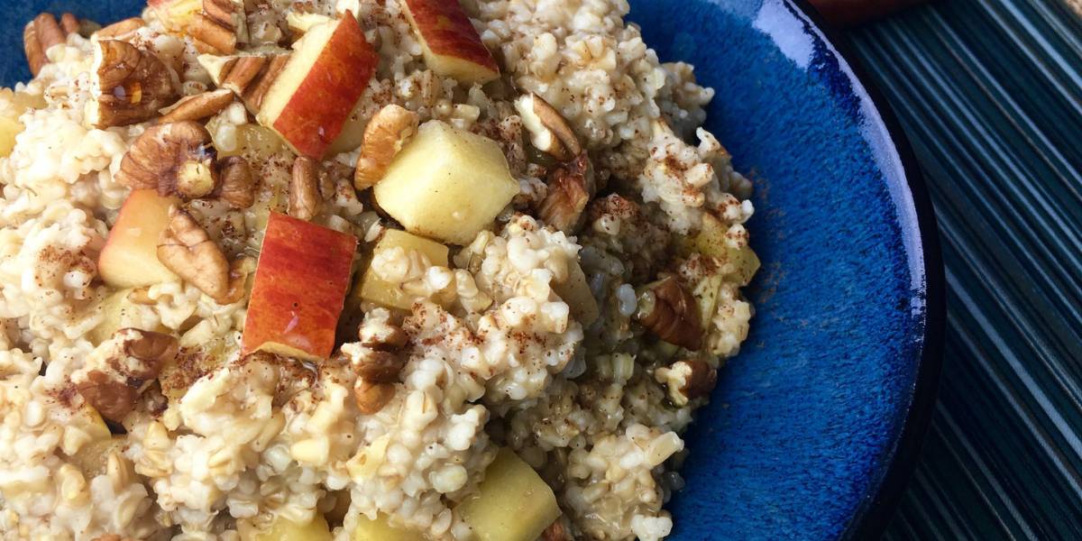 A close-up of a blue bowl filled with oatmeal topped with chopped red apple, pecans, and a sprinkle of cinnamon. The oatmeal has a hearty texture, and the toppings add a colorful and crunchy contrast. The dish is set on a dark, textured surface.