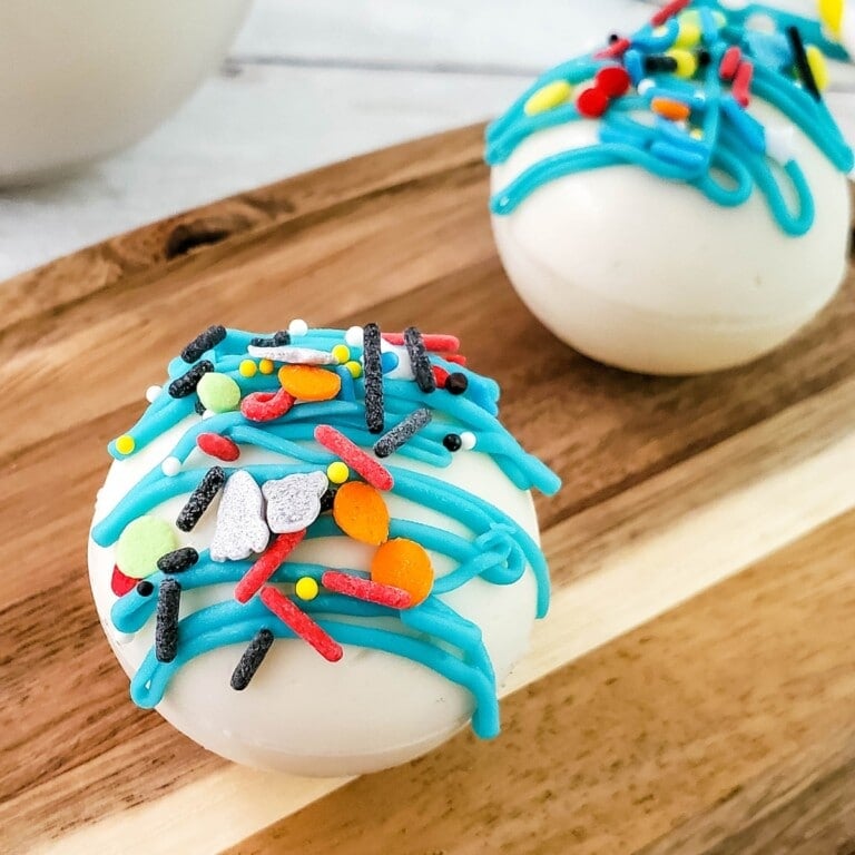 White cocoa bombs with blue frosting and colorful sprinkles on a wooden board.