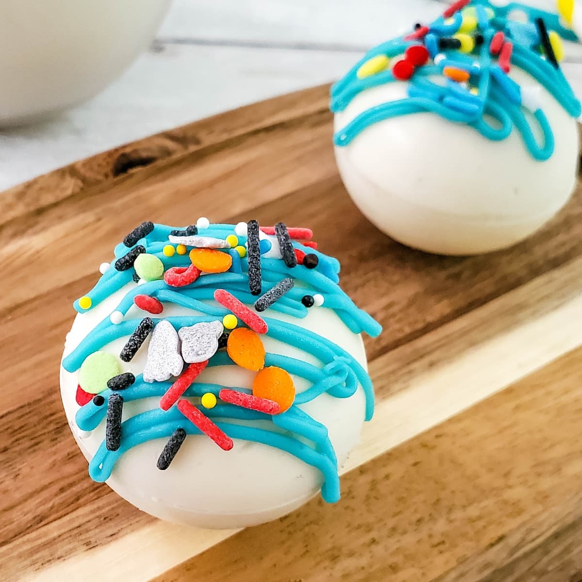 White cocoa bombs with blue frosting and colorful sprinkles on a wooden board.