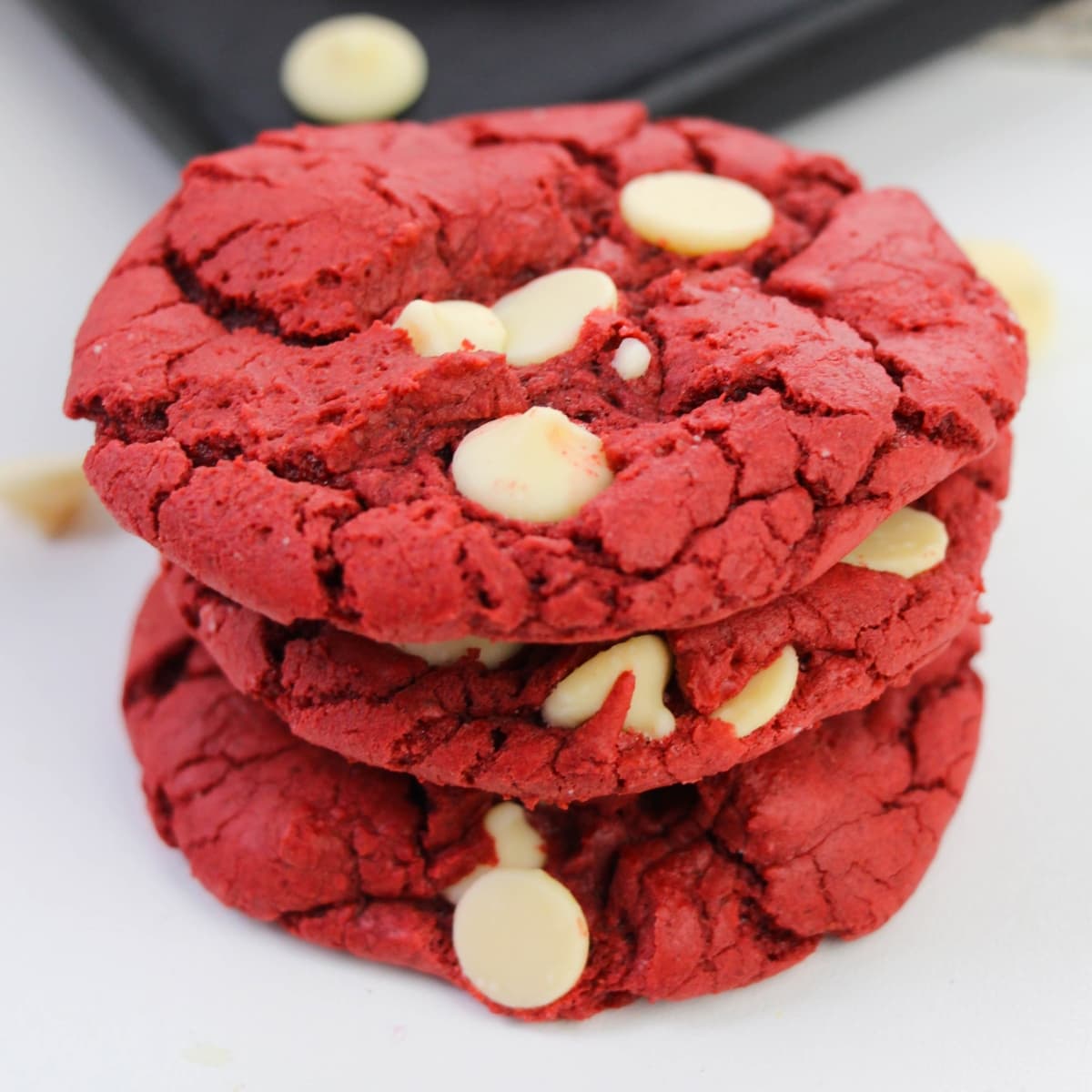 Three red velvet cake mix cookies with white chocolate chips stacked on a white surface.