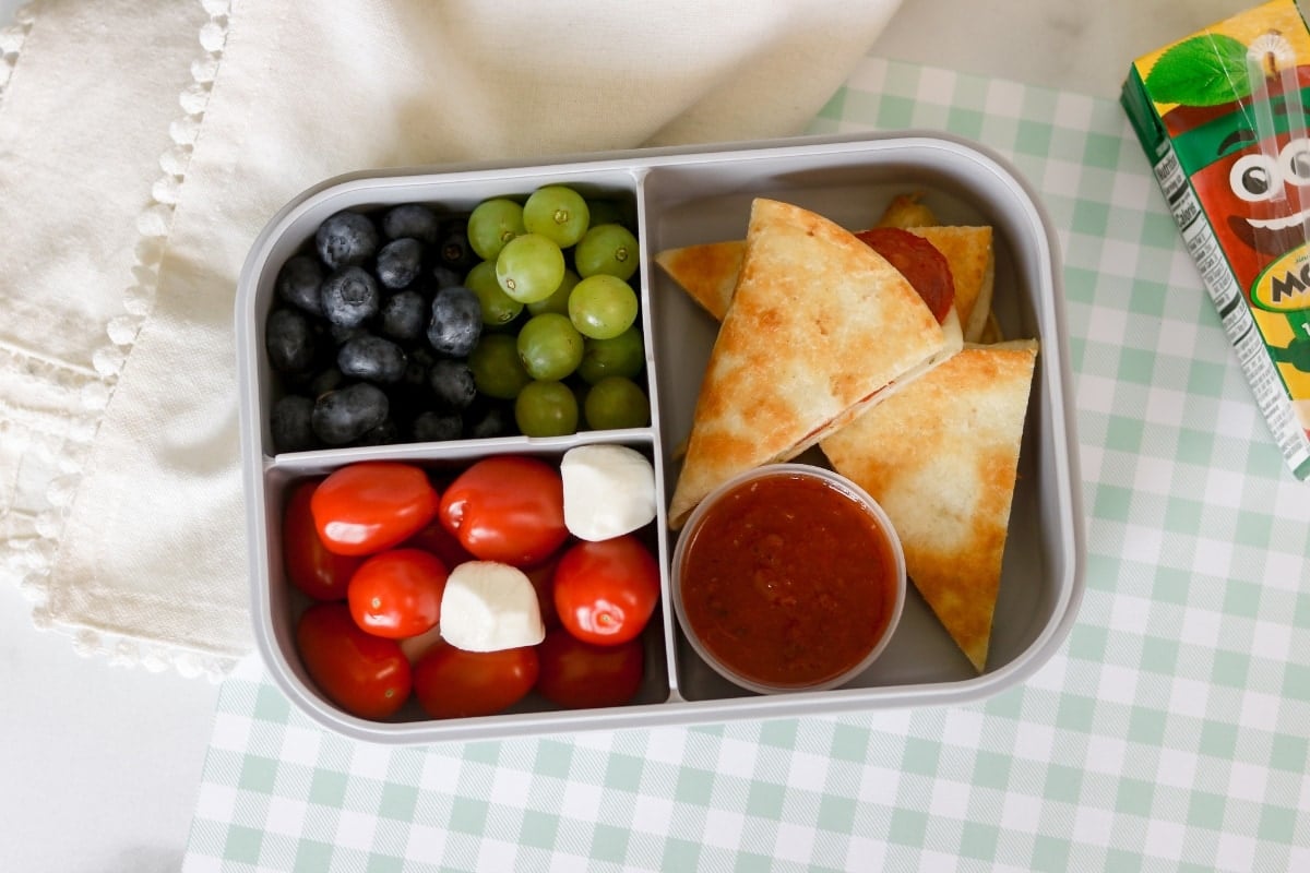 A lunch box with pepperoni pizzadilla with blueberry, green grapes, and tomatoes.