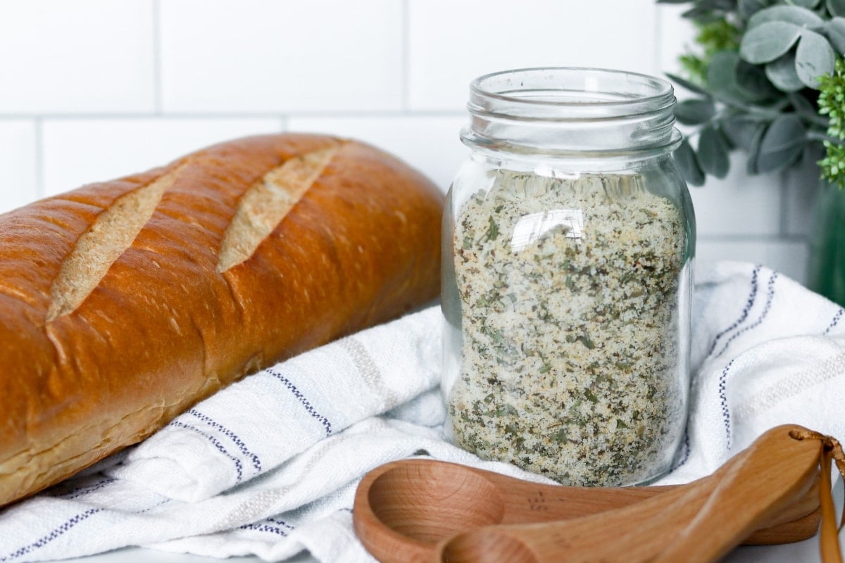 Garlic bread seasoning in a jar with bread and a wooden measuring spoon on the sides.