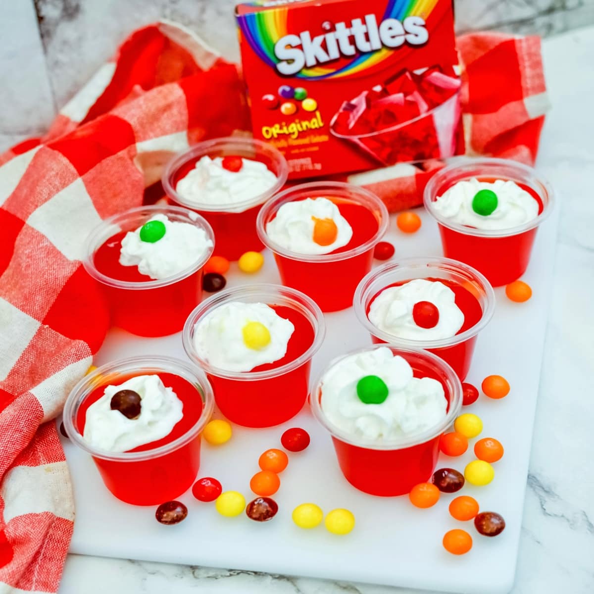 Tray of red jello in plastic condiment cups topped with whipped cream and a skittle.