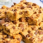 A pile of cookie bars topped with chopped Reese’s peanut butter cups, chopped walnuts, and chocolate chips.