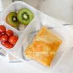 Lunch box with Hot Pockets with a slice of kiwi and cherry tomatoes on the sides.