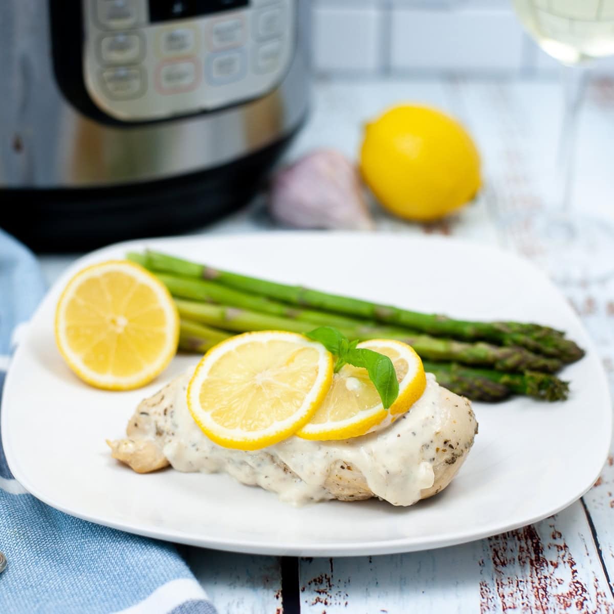 A white plate of breast chicken with creamy sauce and slices of lemon on top.