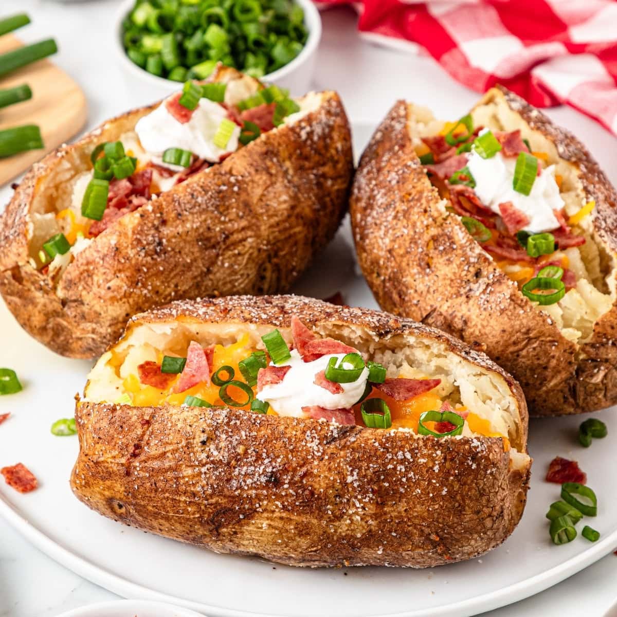 Three baked potatoes on a white plate with cheese, bacon, sour cream, and green onions on top.