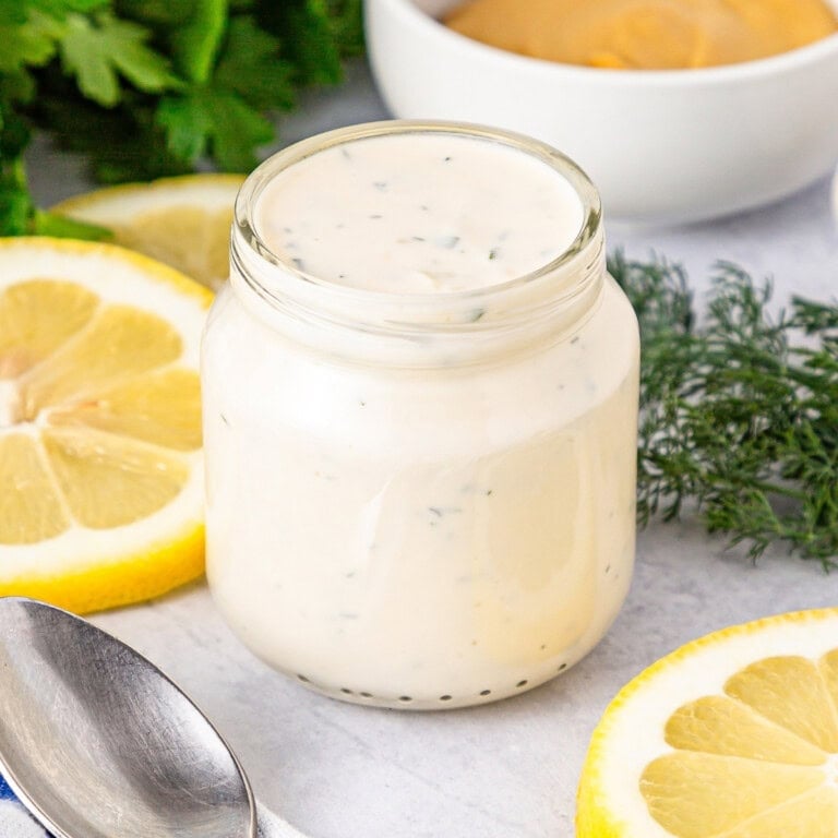 Aioli Sauce in a glass jar with slices of lemon, dill, and parsley in the background.