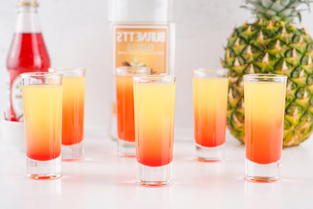 Six Pineapple Upside Down Cake Shots with pineapple, vodka, and grenadine syrup on the background.