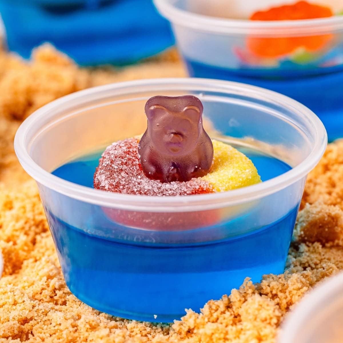 Blue jello shot in a small plastic container topped with a gummy ring and gummy bear.