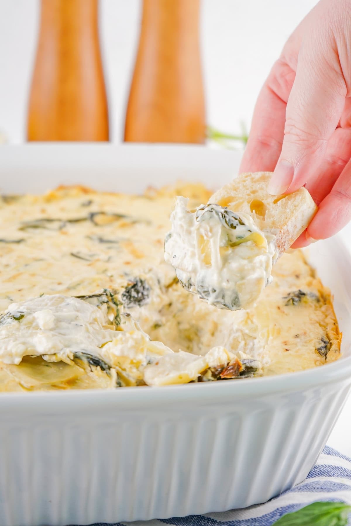 A French bread scooping a spinach artichoke dip in a white casserole.