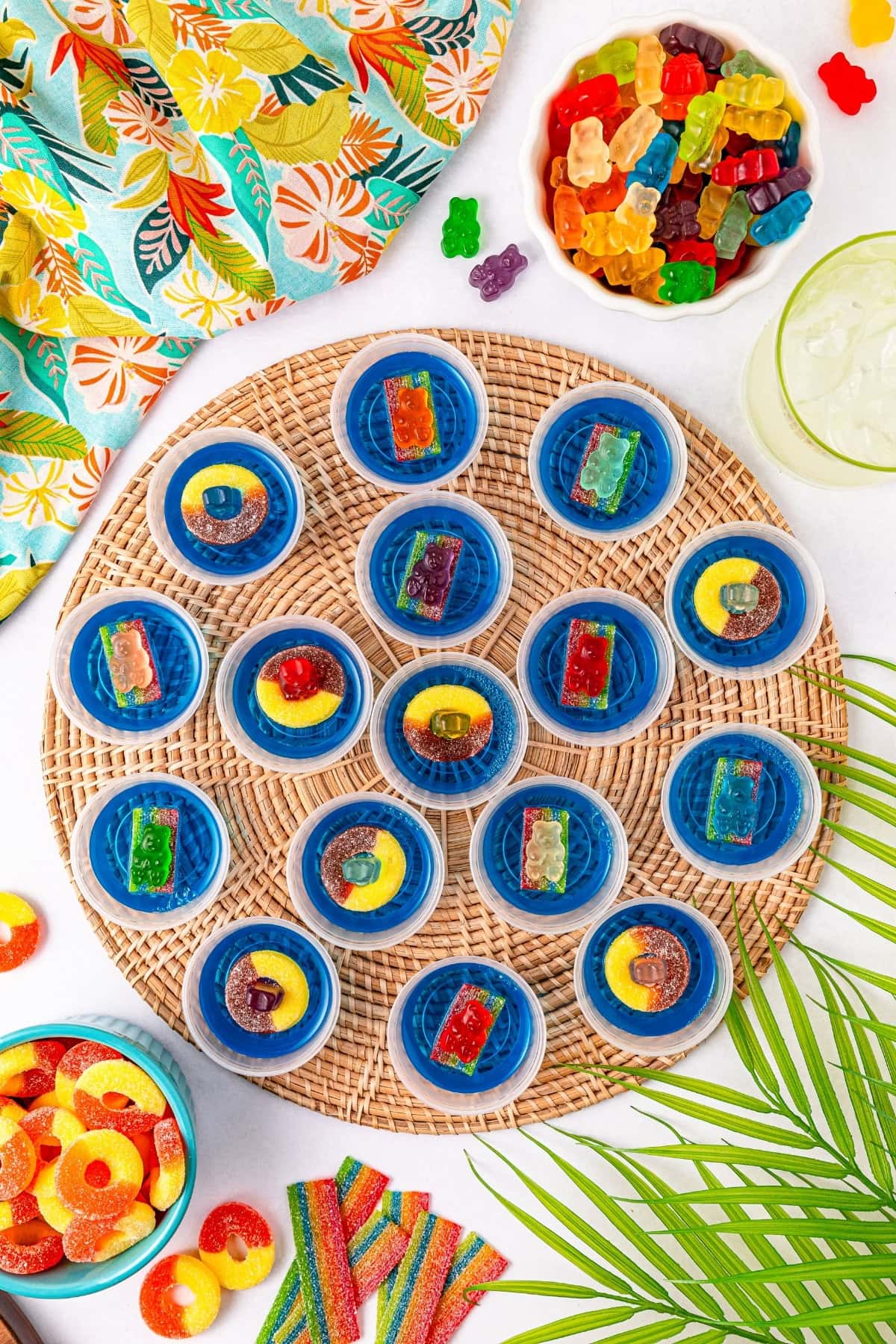A small cup of blue jello shots sitting on a round placemat with gummy bears, gummy rings, and sour rainbow belts in the background.