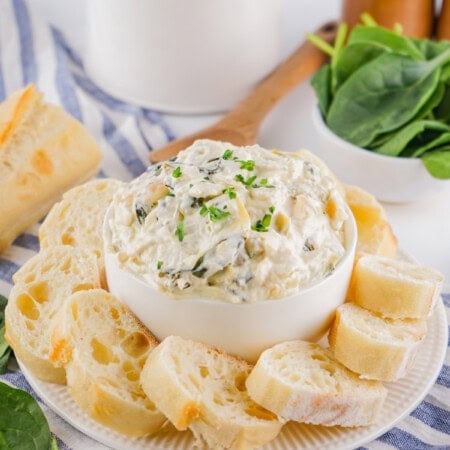 Spinach artichoke dip in a white bowl served with French bread slices on the sides.