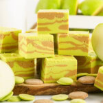 A pile of apple caramel fudges sitting on a wooden board with caramel and green candy melts in the background.