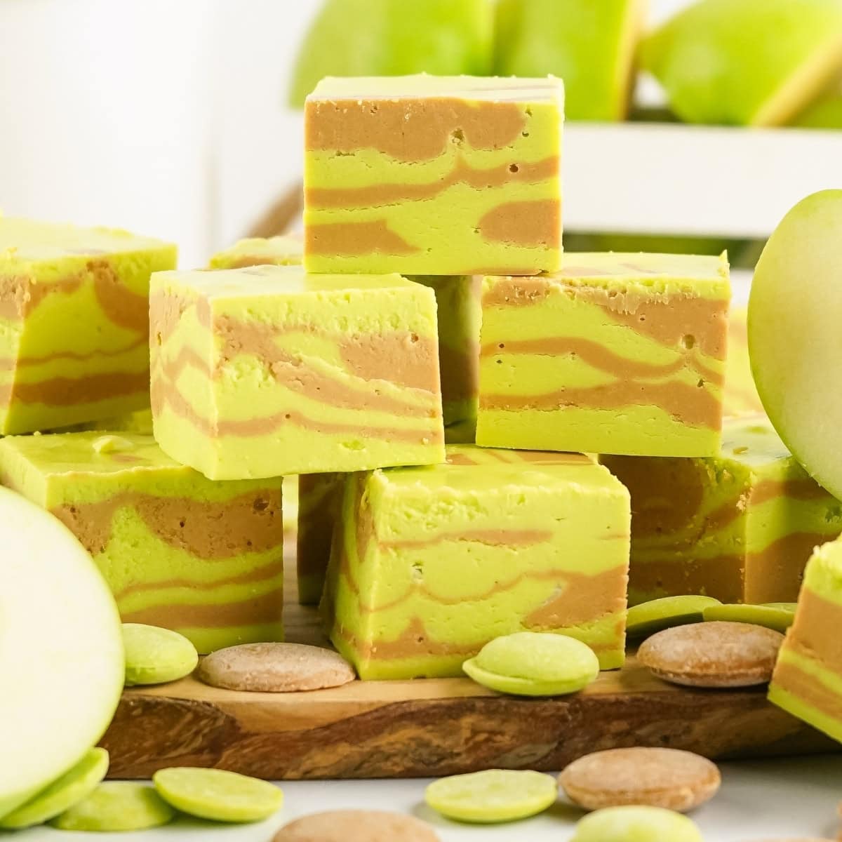 A pile of apple caramel fudges sitting on a wooden board with caramel and green candy melts in the background.