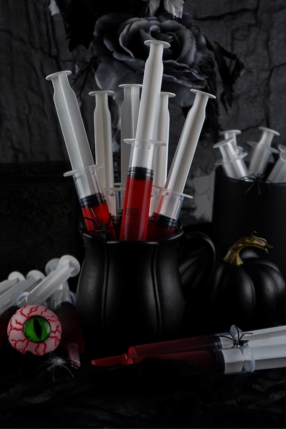 Halloween themed jello shots in syringes sitting in a black cup with Halloween decorations in the background.