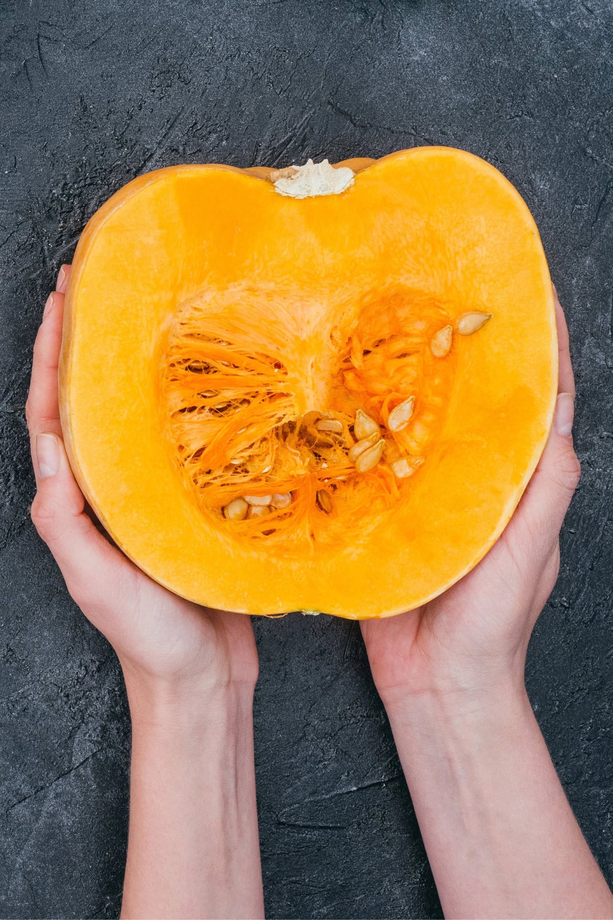 Hands holding a halved squash with seeds and fibers visible against a dark textured background.
