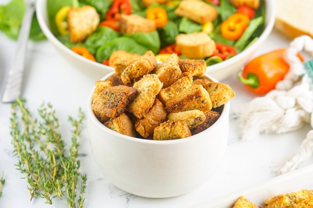 A bowl of seasoned croutons on a table with fresh salad, herbs, and a sliced pepper in the background.