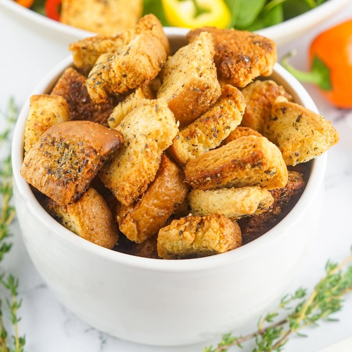 A white bowl filled with seasoned croutons, surrounded by fresh herbs and vegetables on a marble surface.