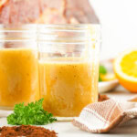 Two jars of ham glaze with parsley and a checkered cloth nearby, plus an orange half and blurred background details.