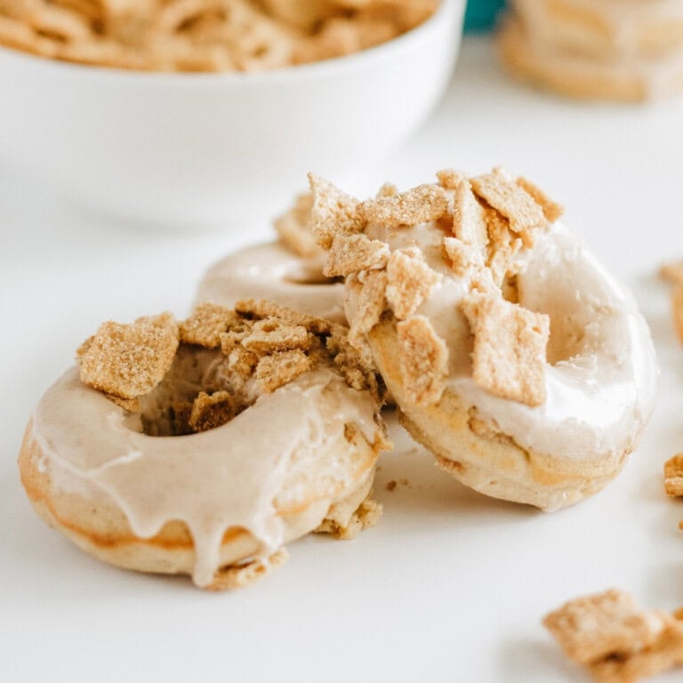 Two glazed donuts topped with crumbled cereal sit on a white surface, with a bowl of additional cereal in the background.