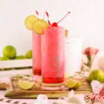 Two tall glasses of pink iced drinks with cherries, lime slices, and straws. Surrounded by ice cubes, limes, and a red cloth.