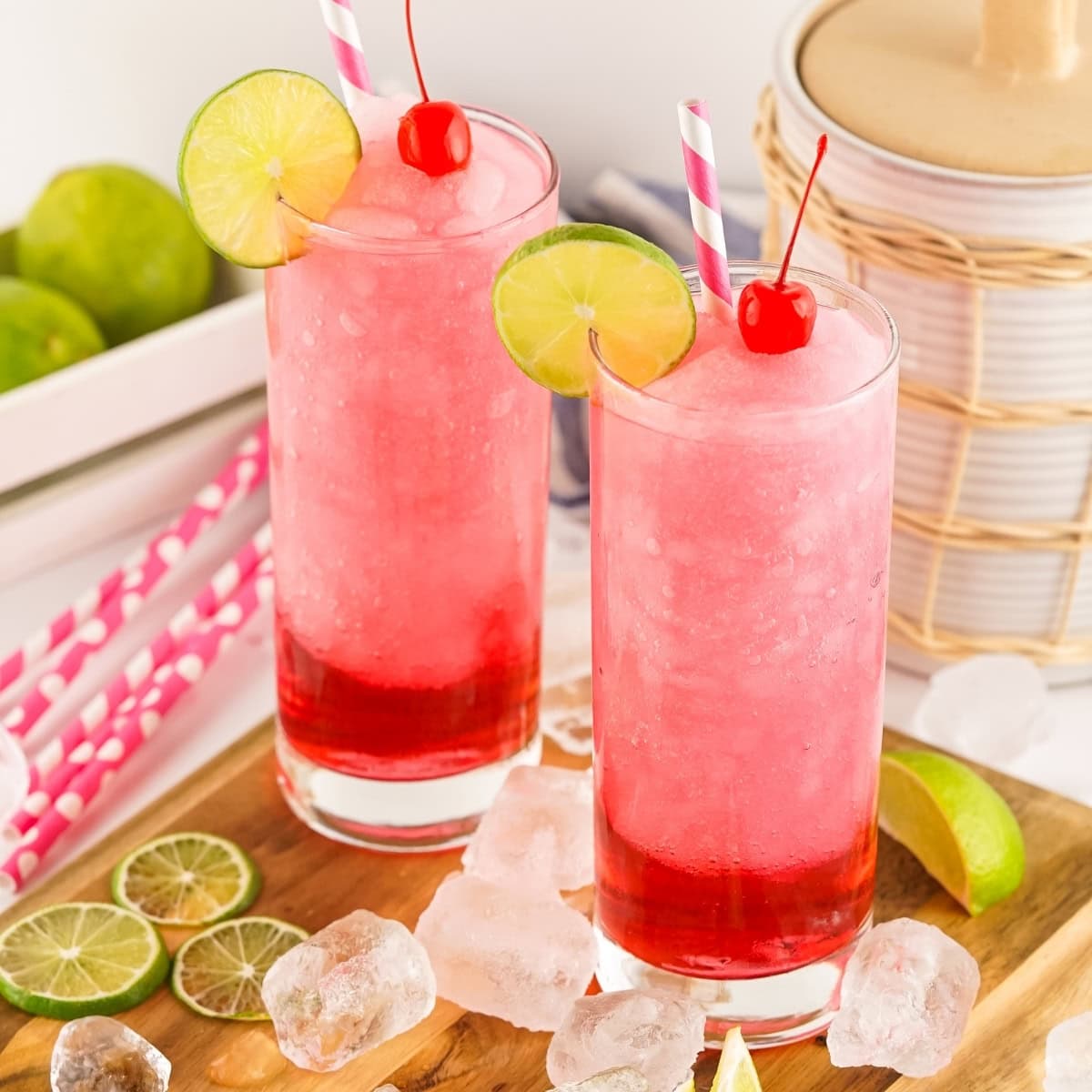 Two tall glasses of pink slushie topped with lime slices and cherries, surrounded by ice cubes and lime wedges on a wooden board.