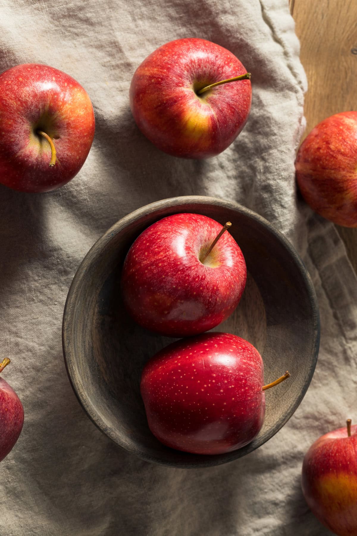 Red apples in a wooden bowl on a cloth, surrounded by more apples.