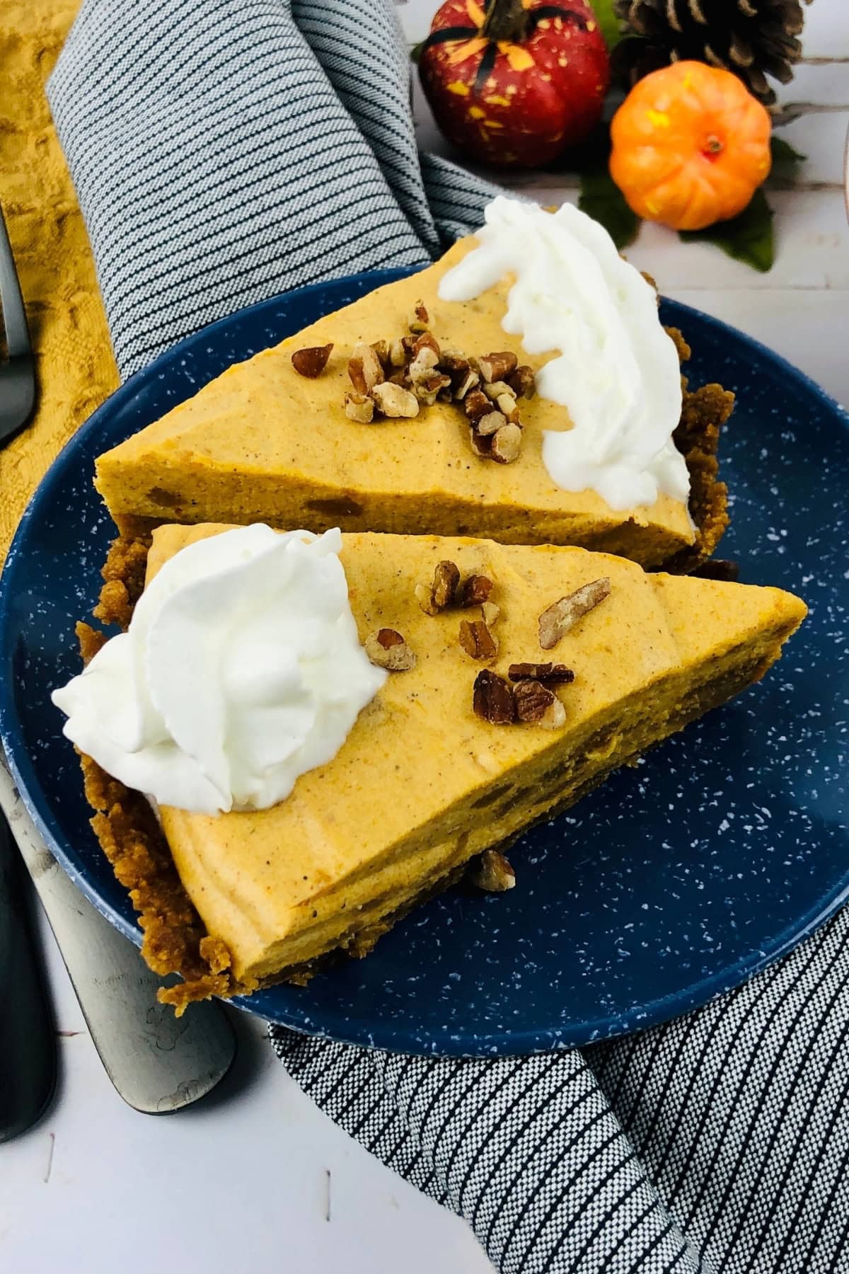 Two slices of pumpkin pie on a blue plate, topped with whipped cream and pecans, with fall decor in the background.