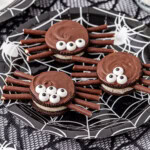 Three spider-themed cookies with candy eyes and chocolate legs on a spiderweb-patterned plate.