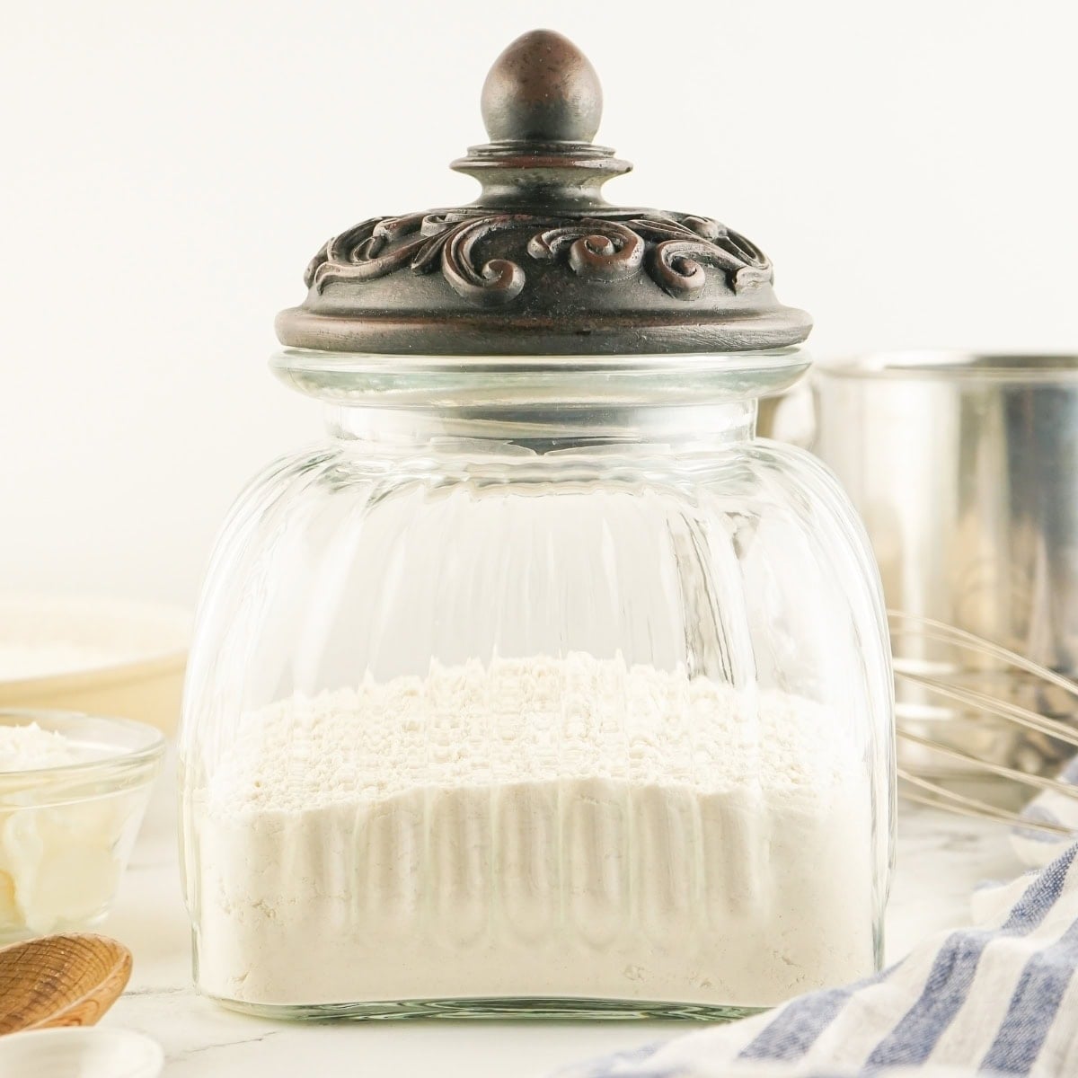 Glass jar with ornate lid filled with biscuit mix on counter.