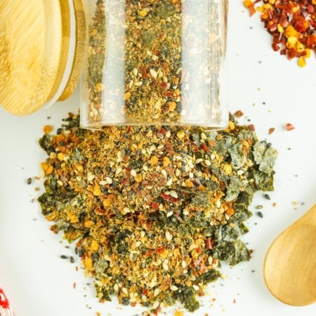 A jar of mixed spices spilled on a white surface, with a wooden spoon and scattered red pepper flakes nearby.