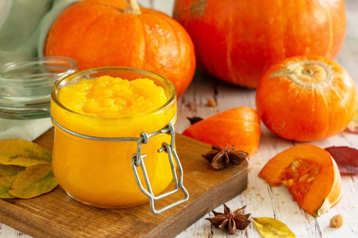 Jar of pumpkin puree on a wooden board, surrounded by whole pumpkins, pumpkin pieces, autumn leaves, and star anise.