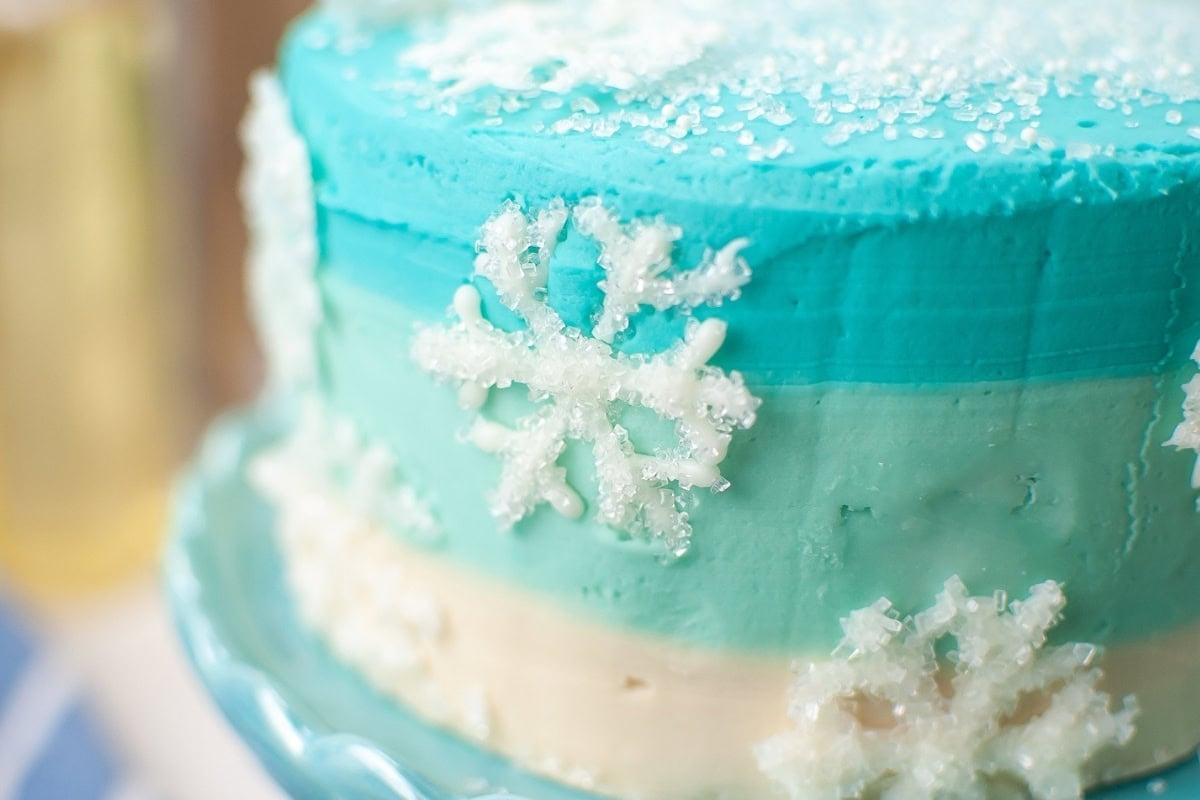 A blue and white cake decorated with snowflakes and sugar crystals on a blue stand.
