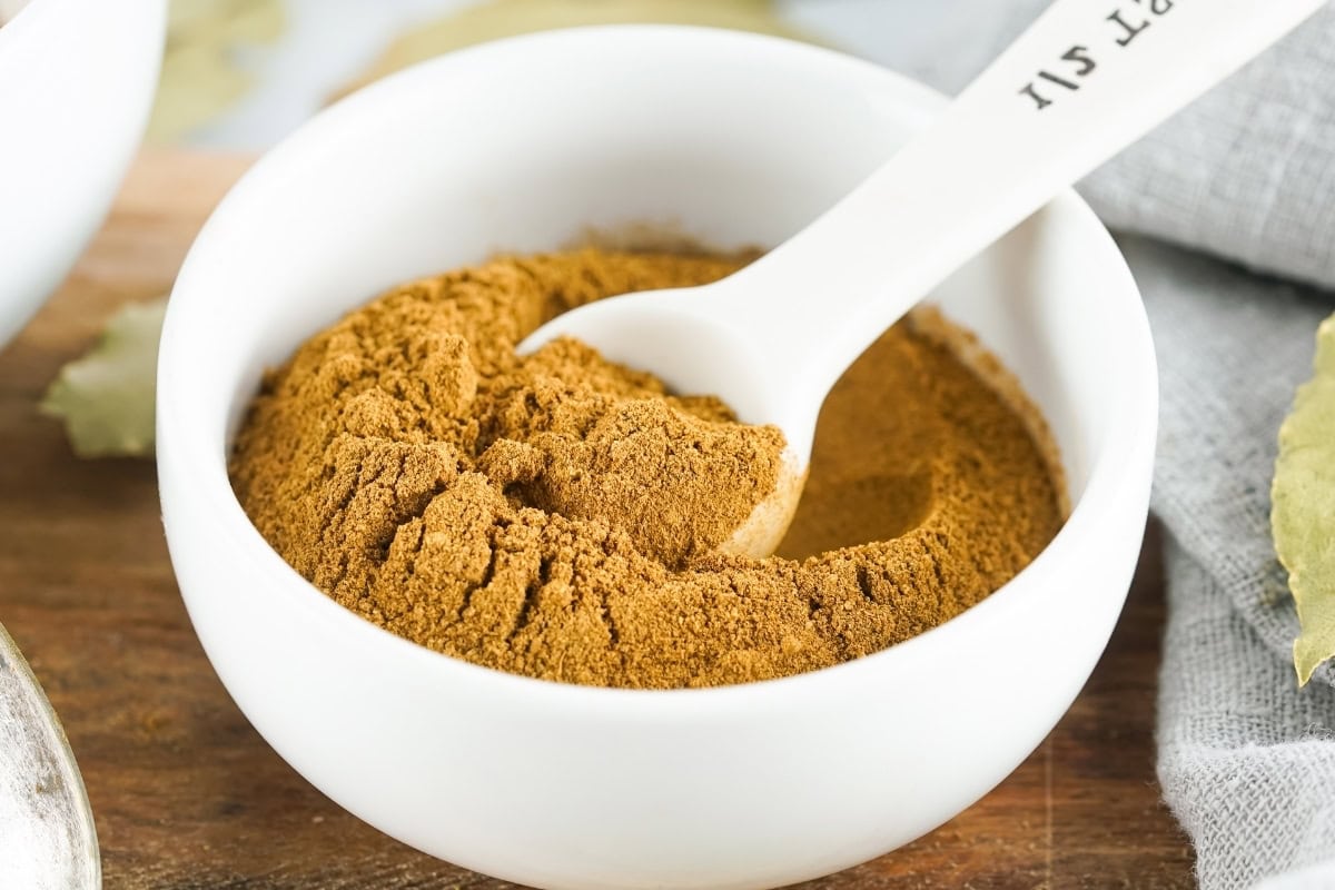 White bowl filled with pumpkin pie spice and a white spoon placed in it, set on a wooden surface with a gray cloth nearby.