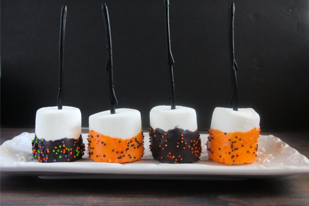 Four marshmallows dipped in orange and black chocolate with sprinkles, on sticks, displayed on a white plate.