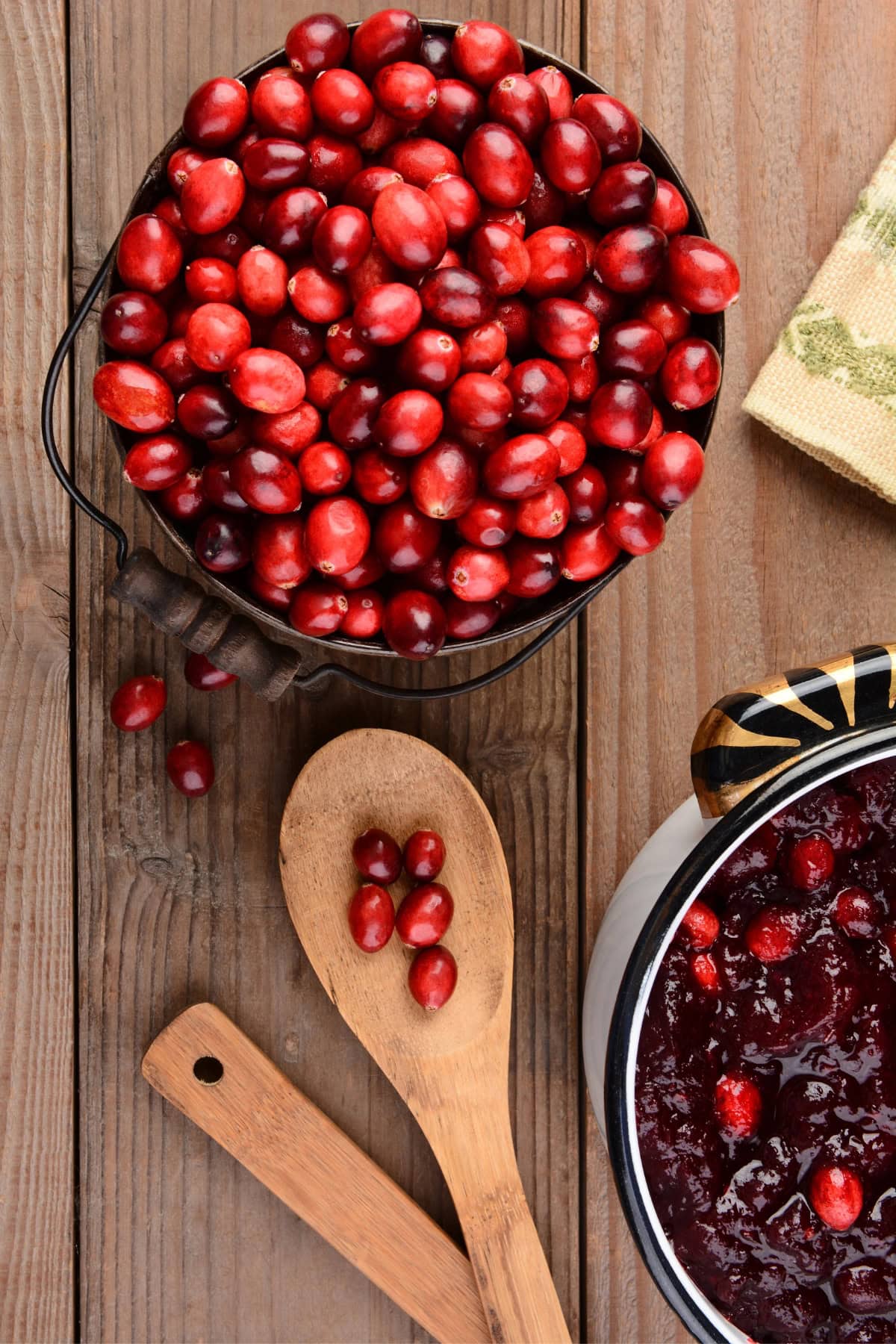 Fresh cranberries in a bowl with a wooden spoon and a pot of cranberry sauce on a wooden table.