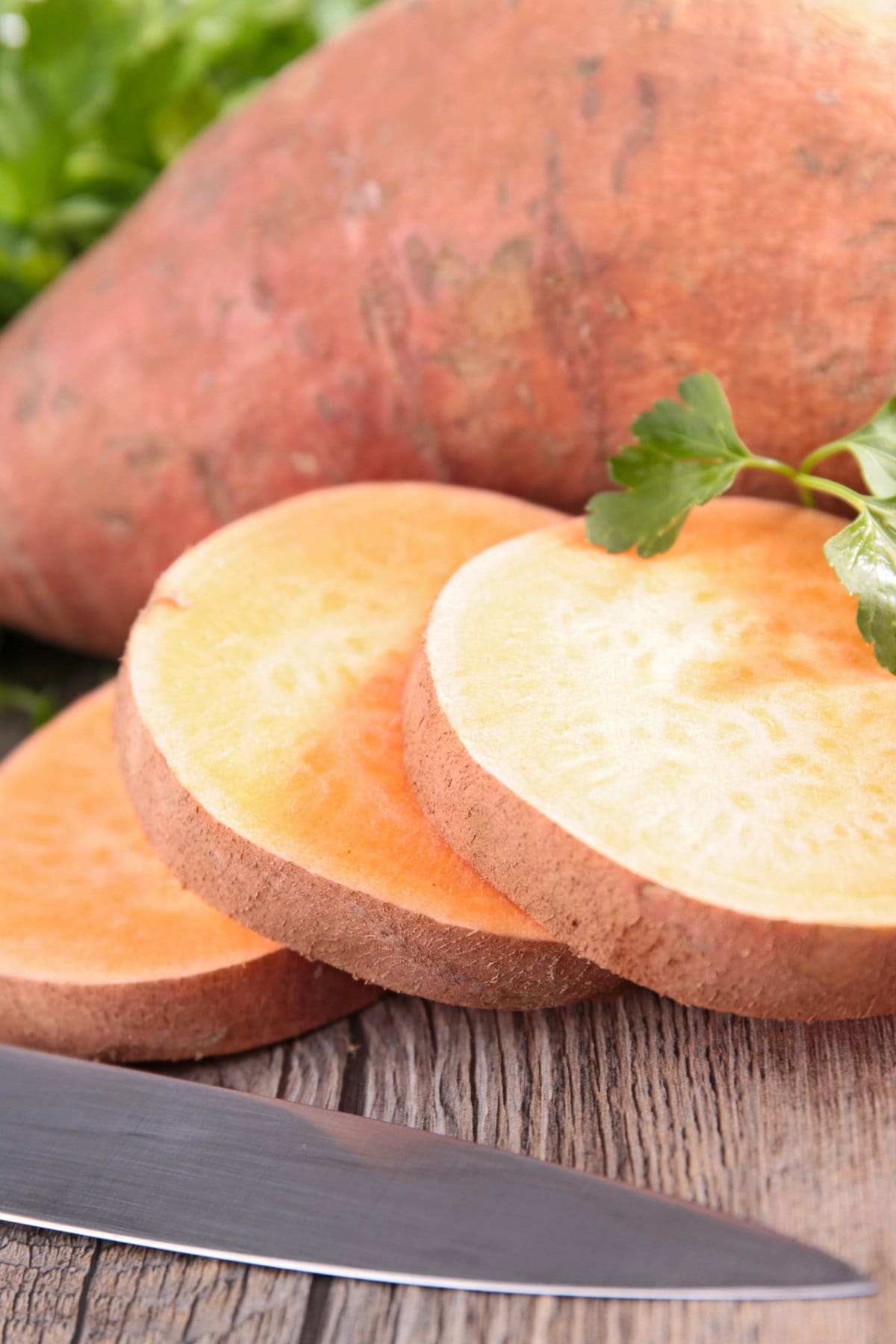 Sliced sweet potato on a wooden board with a knife and parsley.