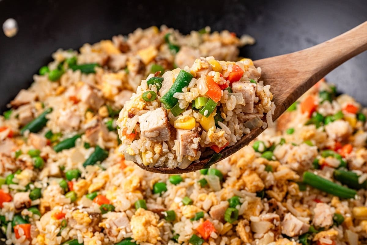 A wooden spoon scoops up colorful vegetable fried rice from a wok.
