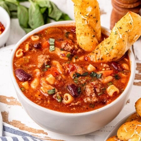 Bowl of pasta e fagioli with breadsticks and herbs on a table.
