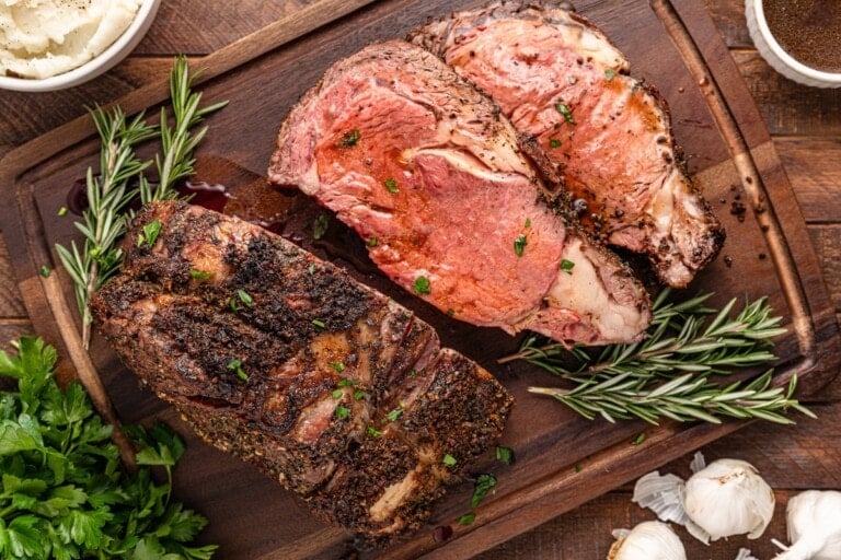 Sliced roast beef on a wooden board with rosemary and garlic.