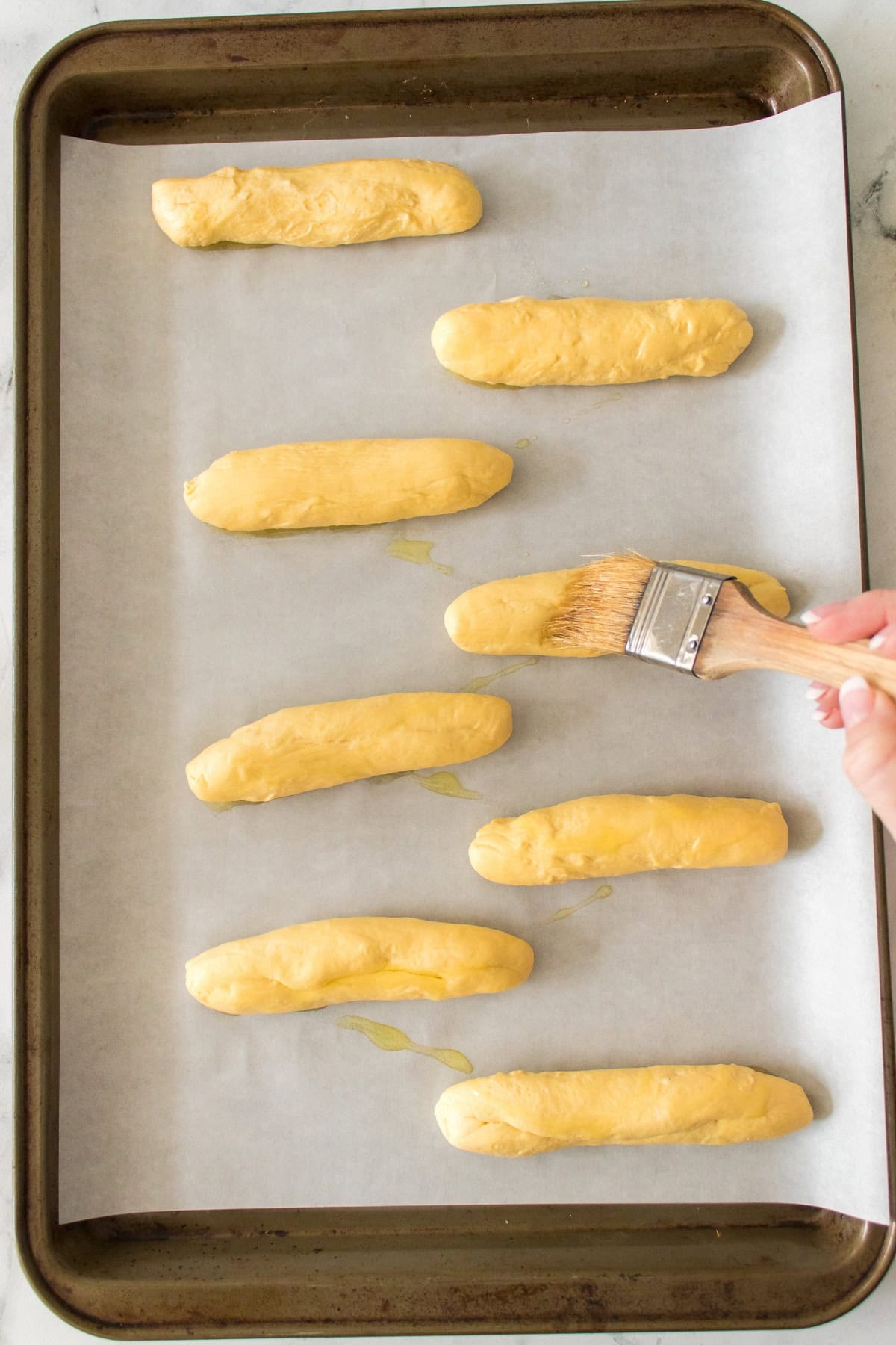 Person brushing breadsticks with oil on a parchment-lined baking sheet.