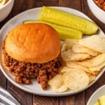 Sloppy joe sandwich with chips and pickle slices on a white plate.