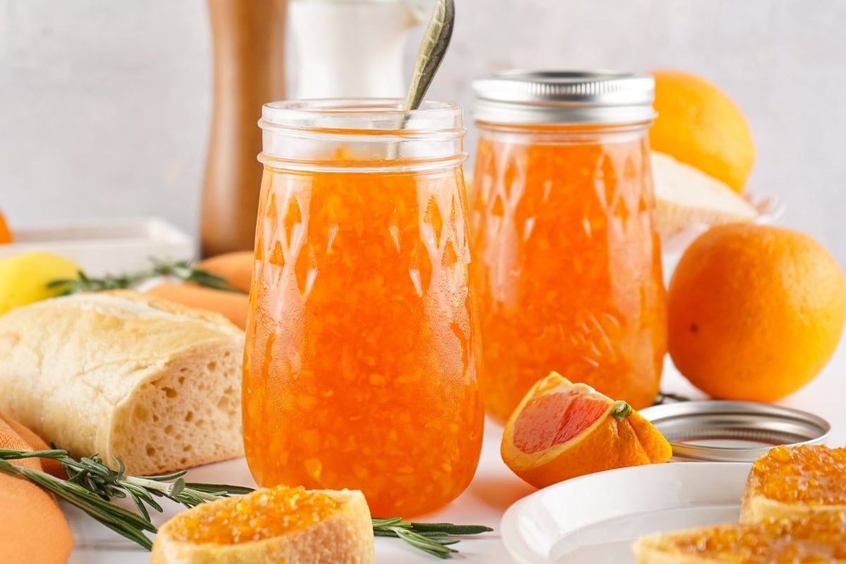 Jars of orange marmalade with bread and oranges on a table.