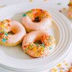Three glazed donuts topped with colorful cereal on a white plate.
