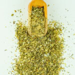Herb and spice mix in a wooden scoop on a white surface.