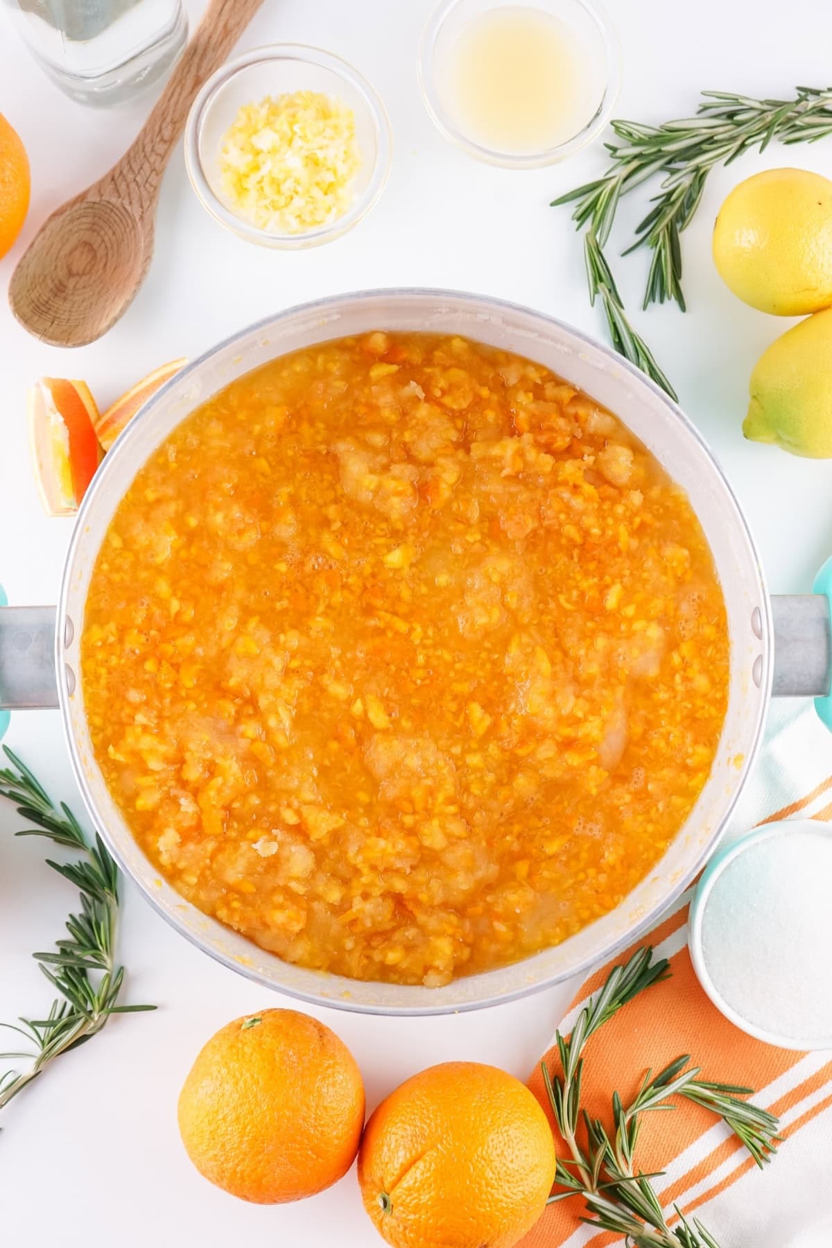 Pot of orange marmalade surrounded by fresh oranges and rosemary sprigs.