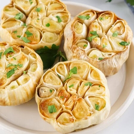 Roasted garlic heads garnished with herbs on a white plate.