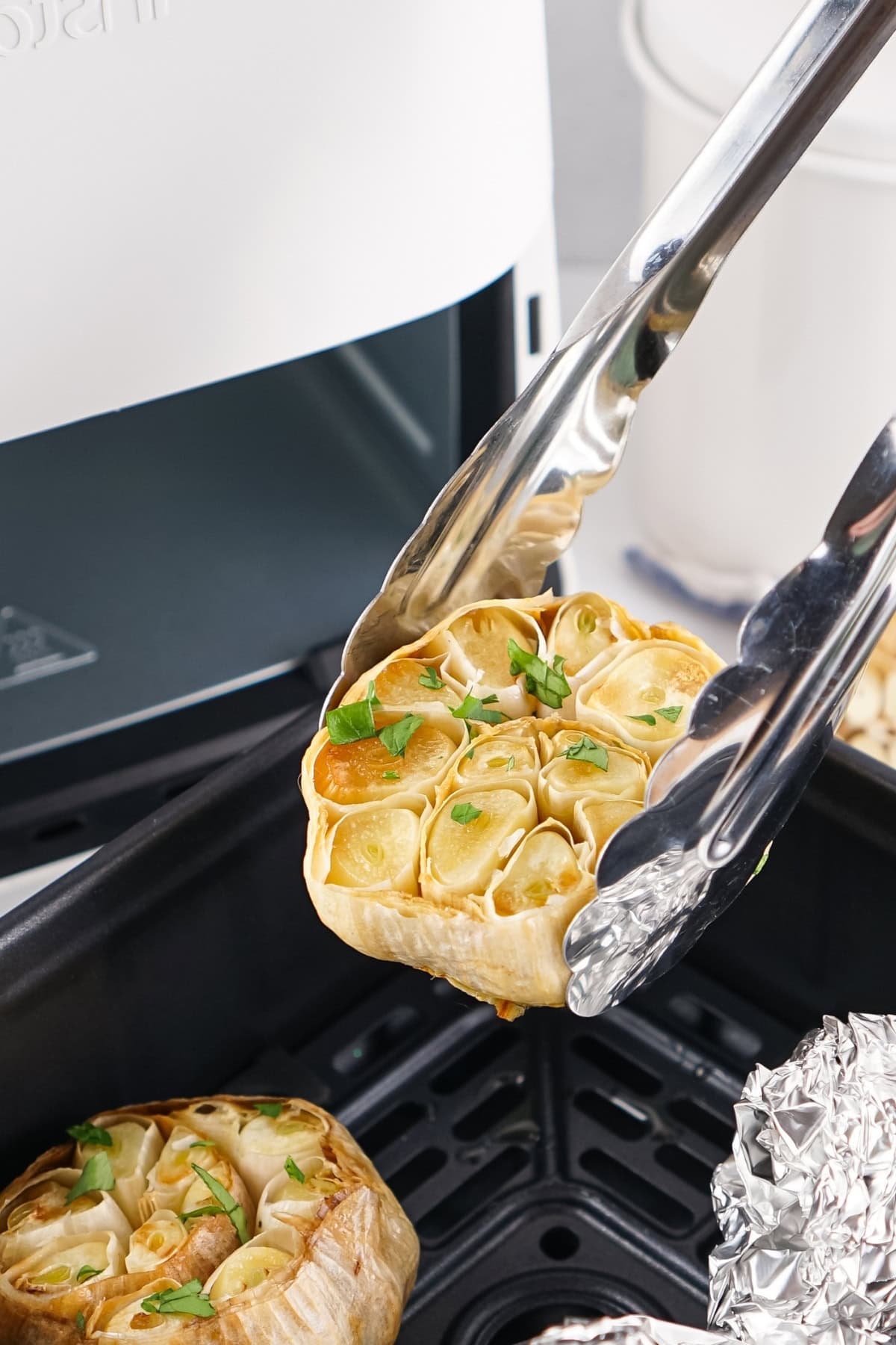 Tongs holding roasted garlic bulb garnished with herbs near an air fryer.
